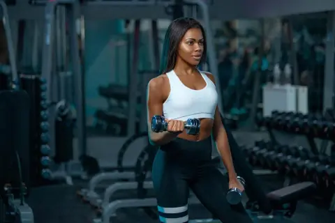 A woman with muscular arms lifts dumbbells in a gym, showcasing her dedication to fitness.