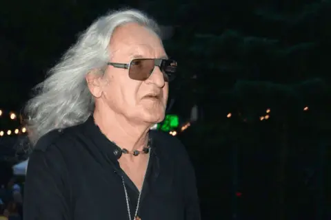 Grzegorz Markowski z długimi siwymi włosami i w okularach przeciwsłonecznych, w czarnej koszuli. W tle widać światła i tłum ludzi.
