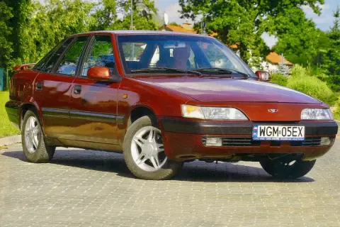 Czerwony Daewoo Espero z lat 90. na tle zieleni i brukowanej drogi.