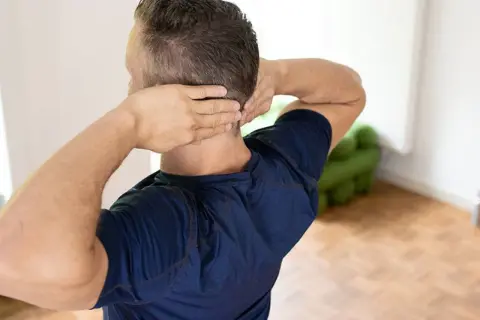 Mann mit Händen im Nacken, der sich fragt: nackenschmerzen wann zum arzt.