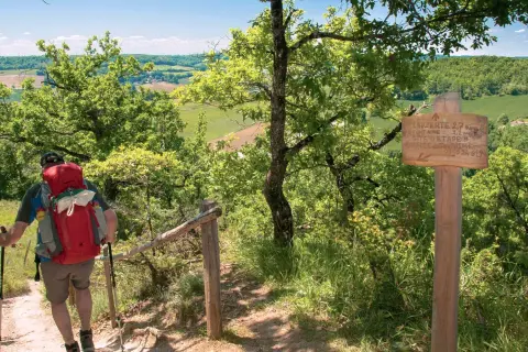 Randonnées Tarn et Garonne : un marcheur avec un sac à dos rouge monte un sentier boisé, dominant un paysage verdoyant. Panneau indicateur de Lauzerte.