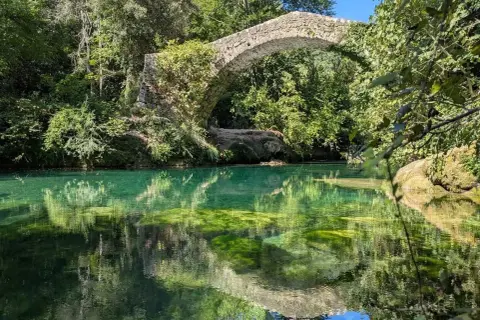 Pont de pierre envahi par la végétation au-dessus des eaux turquoise des gorges de la Siagne, reflétant le ciel et la végétation luxuriante.