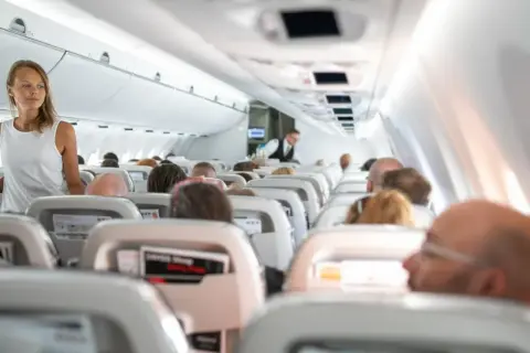 Kobieta w białej bluzce stoi w przejściu samolotu, podczas gdy pasażerowie siedzą w rzędach. Widać lekkie turbulencje w samolocie.