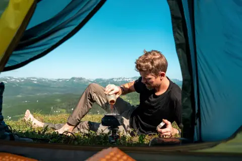 Man häller kaffe i en kastrull vid tältöppningen. En del av packlista camping. Vacker utsikt över fjällen.