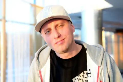 Młody raper w czapce z daszkiem i bluzie z kapturem, z lekkim uśmiechem na twarzy.