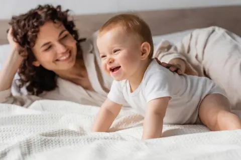 Un bebé de 7 meses gatea en la cama, riendo mientras su madre lo observa con una sonrisa.