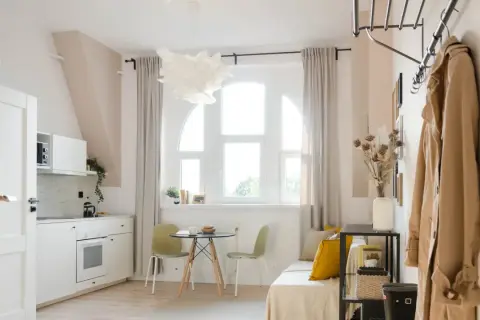Jasne, przytulne mieszkanie idealne na najem okazjonalny. Biała kuchnia, okno z zasłonami, stolik z krzesłami i wygodna sofa.