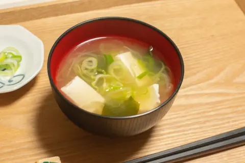 Una ciotola di brodo di miso fumante con tofu e cipollotti, accompagnata da una piccola ciotola di cipollotti affettati e bacchette.