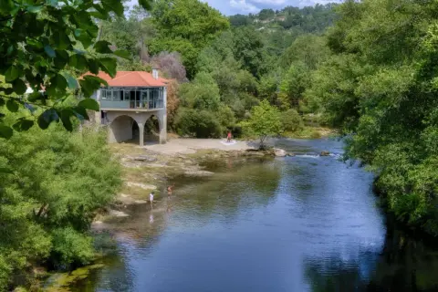 Disfrutando del sol y el agua en la playa fluvial Mondariz. Un edificio con arcos y tejado rojo se alza junto al río, donde unas personas se refrescan.