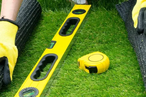 Manos enguantadas preparan terreno para césped artificial. Nivel y cinta métrica listos para el proyecto.