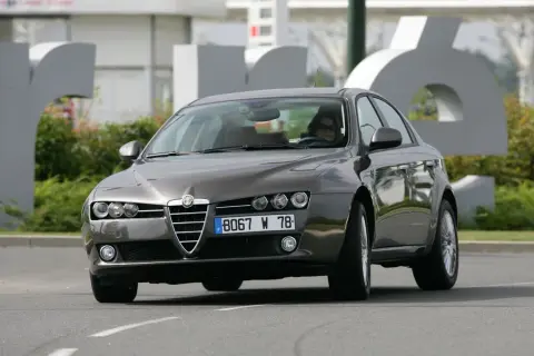Eleganckie Alfa Romeo 159 2.4 JTDm w kolorze grafitowym, z kobietą za kierownicą, jedzie po drodze.