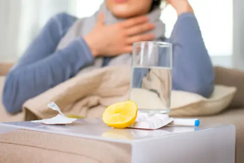 Kobieta z chrypką, na stoliku szklanka wody, cytryna, tabletki i termometr.