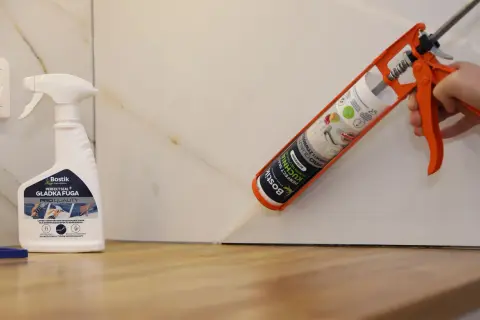 Aplikacja silikonu do blatu kuchennego. Pistolet z kartuszem Bostik Perfect Seal Kuchnia aplikuje masę uszczelniającą. Obok stoi butelka preparatu do wygładzania fug.