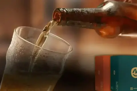 Versando una hefe birra dorata in un bicchiere bagnato. Sullo sfondo, due scatole colorate con il logo "m".