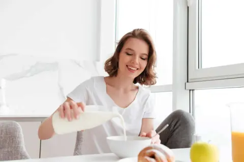 Uśmiechnięta dziewczyna nalewa mleko do miski, dbając o swoją masę kostną. Na stole croissant i jabłko.