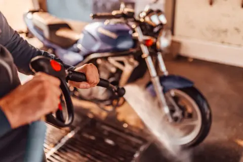 Persona limpiando una moto azul con una hidrolavadora. El agua salpica sobre la rueda delantera.