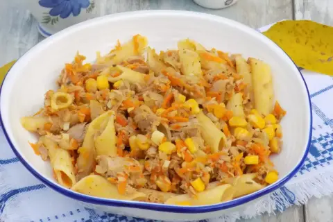 Penne z tuńczykiem, kukurydzą i startą marchewką w białej misce na niebiesko-białej serwetce.