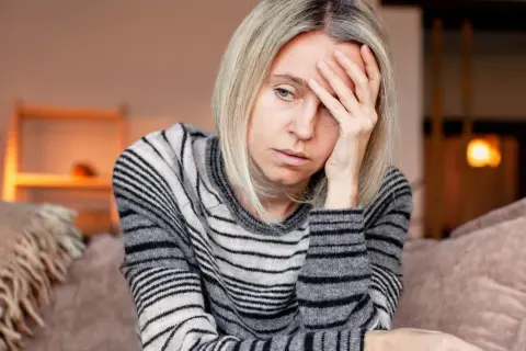 Kobieta z blond włosami, w swetrze w paski, trzyma dłoń na czole, wyraźnie cierpiąc z powodu silnego bólu głowy.