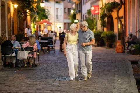 Ein älteres Paar spaziert abends durch eine Gasse in Freiburg. Was kann man in Freiburg machen? Hier: gemütlich essen gehen und die Atmosphäre genießen.