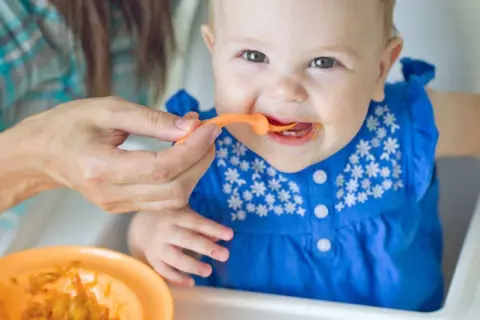 Ein Baby in einem blauen Kleid erhält Brei vom Löffel. Die Augen des Babys strahlen, ein Zeichen für die ersten reifezeichen baby.