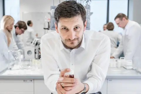 Mężczyzna w białej koszuli trzyma fiolkę z płynem, w tle laboratorium, gdzie pracują inni naukowcy. To kluczowe xtpl akcje.