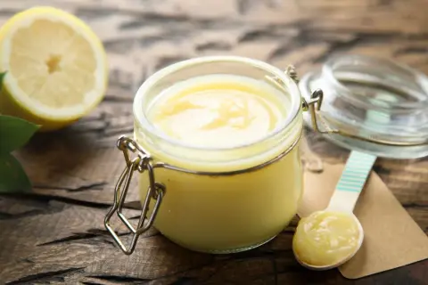 Un pot de lemon curd onctueux, une moitié de citron et des feuilles vertes sur fond de bois. Une cuillère en bois est prête à déguster.