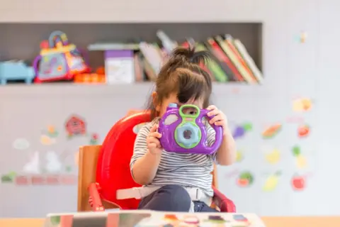 A young child in a striped shirt sits in a red high chair, holding a purple toy camera to their face.