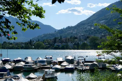 Båtar täckta med presenningar vid en brygga vid Lago Maggiore. Berg och en stad syns i bakgrunden.