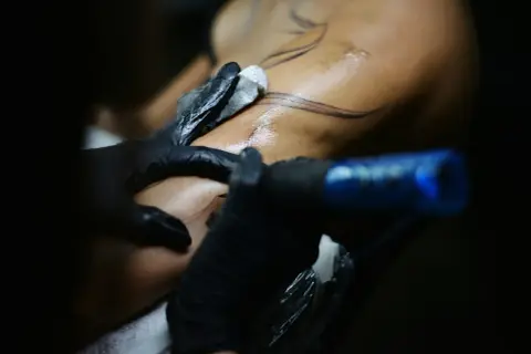 Detailaufnahme: Tätowierer mit schwarzen Handschuhen arbeitet an einem Arm. Feine Linien für blackwork tattoos werden auf die Haut gebracht.