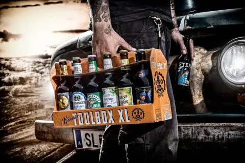 Mężczyzna z tatuażami niesie skrzynkę "TOOLBOX XL" z różnymi butelkami piwa. Idealne połączenie: jakie butelki do jakich skrzynek.