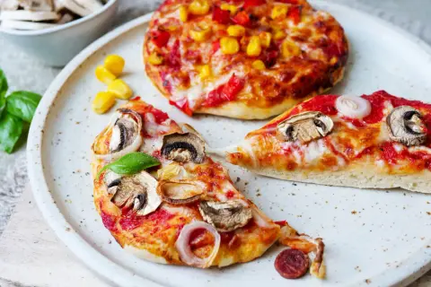 Zwei kleine Pizzen, eine mit Pilzen und Zwiebeln, die andere mit Mais und Paprika, warten darauf, aufgebacken zu werden.