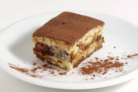 Une part de tiramisu, avec sa traduction française, est saupoudrée de cacao sur une assiette blanche.
