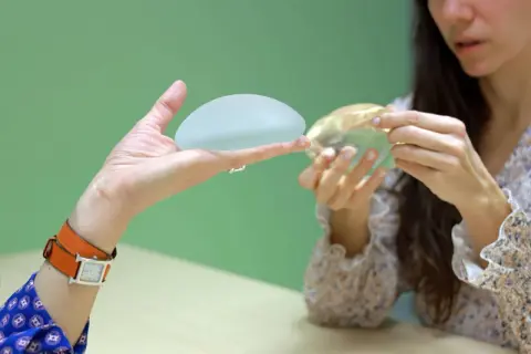 Une main présente un implant mammaire, tandis qu'une autre femme examine un implant différent. Discussion sur la réduction mammaire.