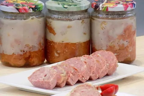 Trzy słoiki domowej kiełbasy w galarecie, obok pokrojona kiełbasa na talerzyku.