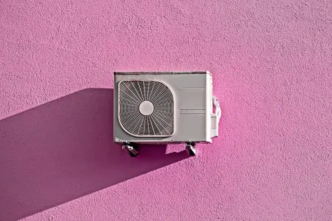 Unità esterna di condizionatore appesa a un muro rosa acceso, con ombra proiettata. Pensando allo smaltimento condizionatori.