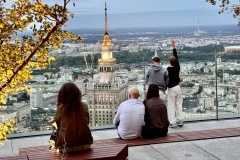 Ludzie podziwiają panoramę Warszawy z Pałacem Kultury i Nauki. Widać wiele warszawskich wież i budynków.