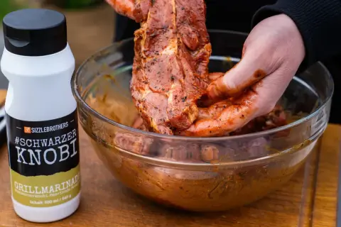 Fleisch wird mit einer würzigen Grillmarinade in einer Schüssel vermischt. Die Flasche "#Schwarzer Knobi" zeigt, wie einfach es ist, Grillmarinade selber machen.