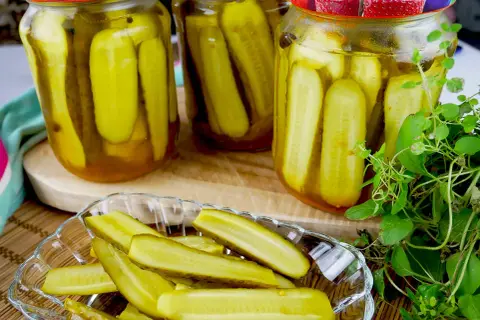 Słoiki pełne ogórków z curry, gotowe do słoików. Na talerzyku pokrojone ogórki, obok świeża zielenina.