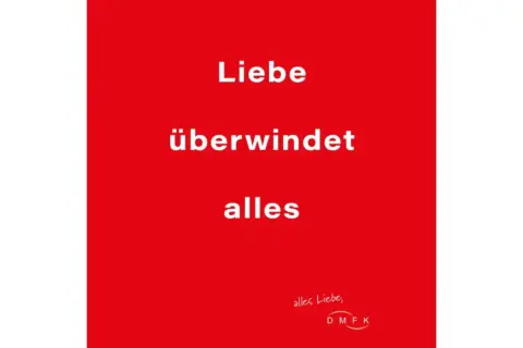 Rode achtergrond met witte tekst: "Liebe überwindet alles". Een boodschap dat liefde overwint alles, met een kleine handgeschreven tekst en logo rechtsonder.