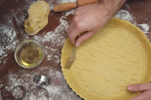 Mani che preparano una pasta frolla morbida, bucherellandola con una forchetta in una tortiera. Farina e ingredienti sparsi sul tavolo.