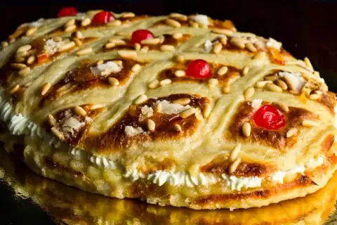 Deliciosa coca de San Juan rellena de crema, decorada con piñones, cerezas rojas y azúcar.