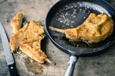 Ein paniertes Kotelett braten in der Pfanne. Daneben liegen bereits geschnittene Stücke auf einem Holzbrett, daneben ein Messer.