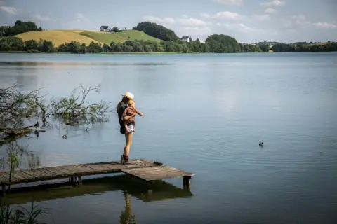Kobieta z dzieckiem na rękach na pomoście, podziwiająca krajobraz jeziora i wzgórz. Szwajcaria Kaszubska atrakcje w pełnej krasie.