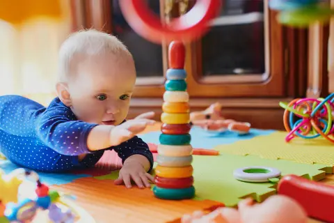 Zaintrygowany 6 miesięczne dziecko sięga do kolorowej wieży z kółek, otoczony zabawkami na macie.
