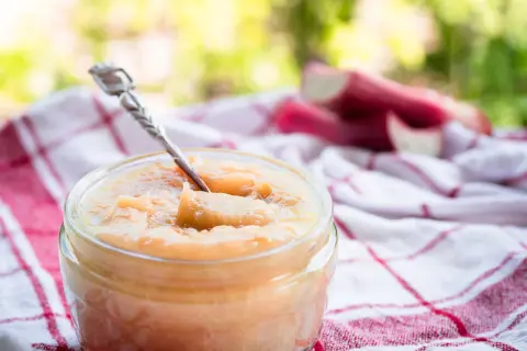 Un pot de compote pomme rhubarbe, avec une cuillère en argent, sur un torchon à carreaux rouges et blancs. Des tiges de rhubarbe sont visibles en arrière-plan.