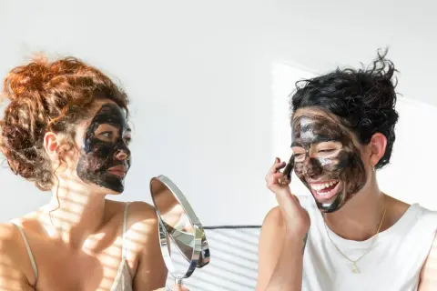Dos mujeres con mascarilla negra en el rostro, una se mira en un espejo, buscando quitar espinillas negras grandes.