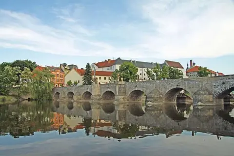 Historische Steinbrücke über den Fluss in Písek, Tschechien, mit Spiegelung im Wasser und bunten Häusern im Hintergrund.