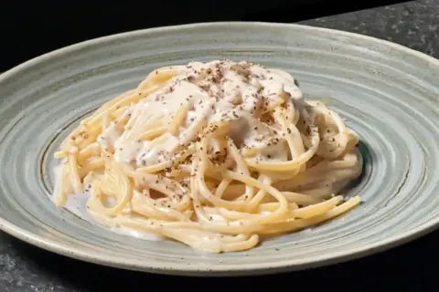 Un plat de tonnarelli cacio e pepe crémeux, saupoudré de poivre noir fraîchement moulu, servi dans une assiette texturée.