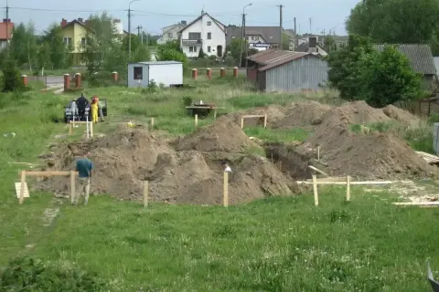 Prace budowlane w toku: kopanie wykopy pod fundamenty, ziemia usypana w kopce. W tle domy i pracownicy.