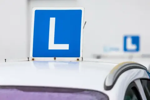 Tablica "L" na dachu białego samochodu. Zbliża się egzamin na prawo jazdy, a ten znak symbolizuje naukę jazdy.